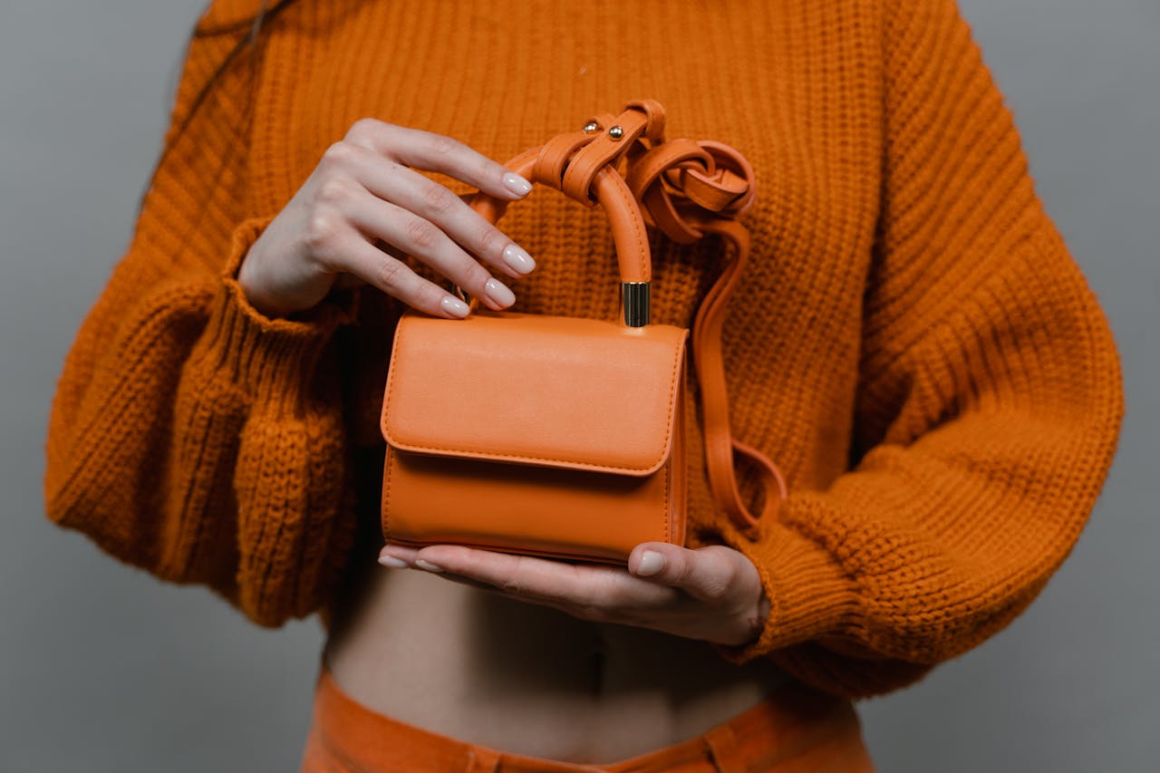 gallery-01 Woman holding an orange leather mini handbag against an orange sweater in a studio setting.